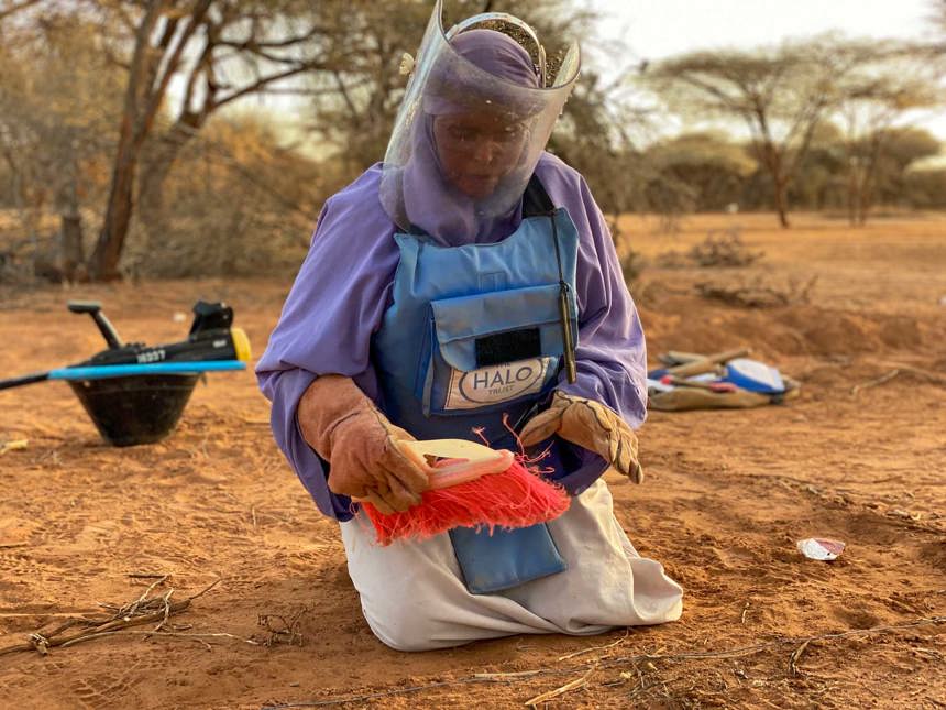 A female deminer wearing PPE wears gloves and holds a brush while kneeling on the soil