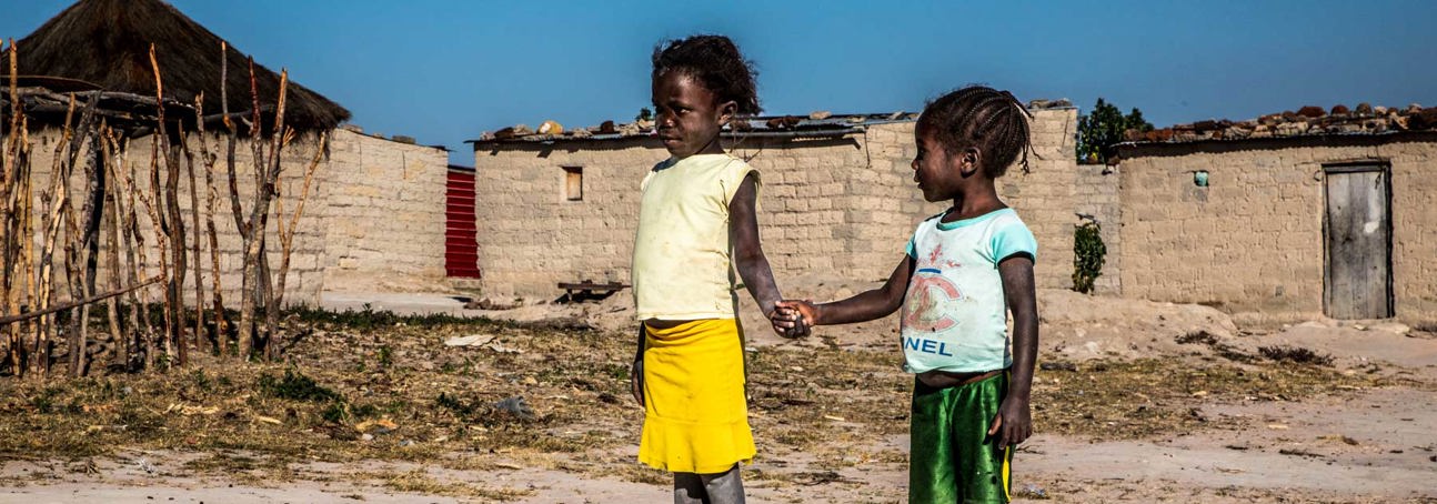 Two children hold hands outside of a house in the village of Lusserei Village, Angola