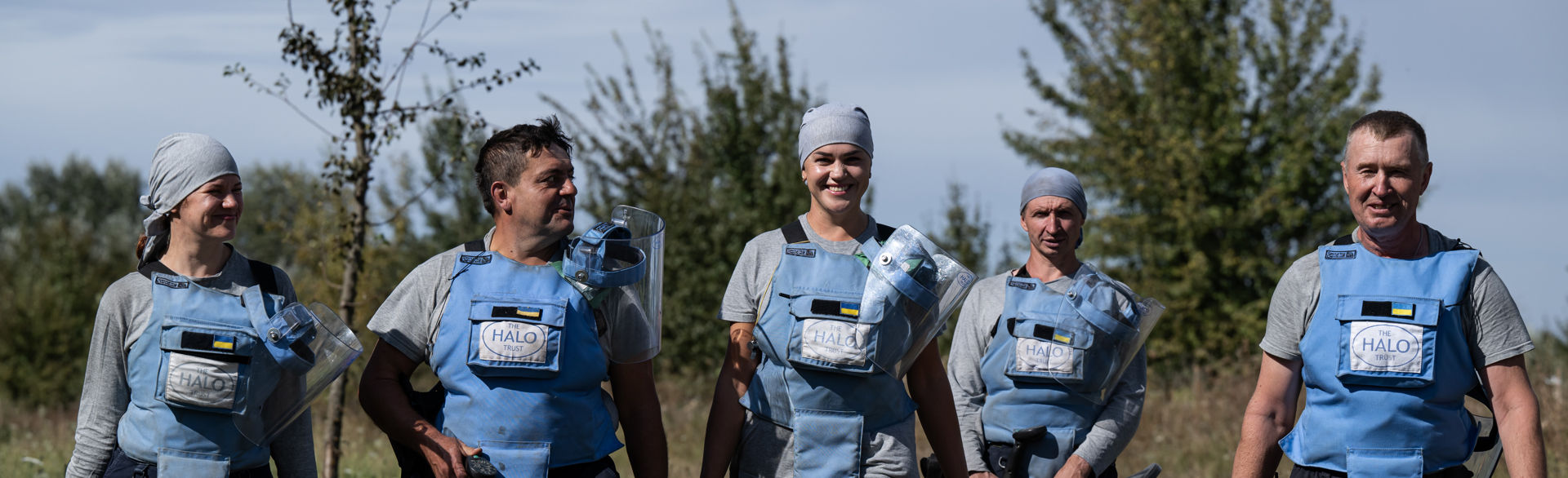 Five smiling deminers in blue HALO vests walk towards the camera in a line.