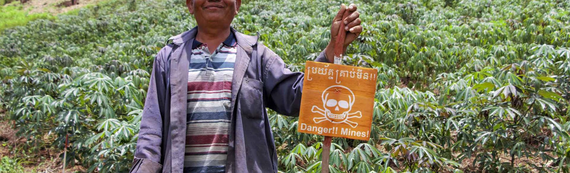 Pork Chei stands next to a mine warning sign in a Cambodian crop field