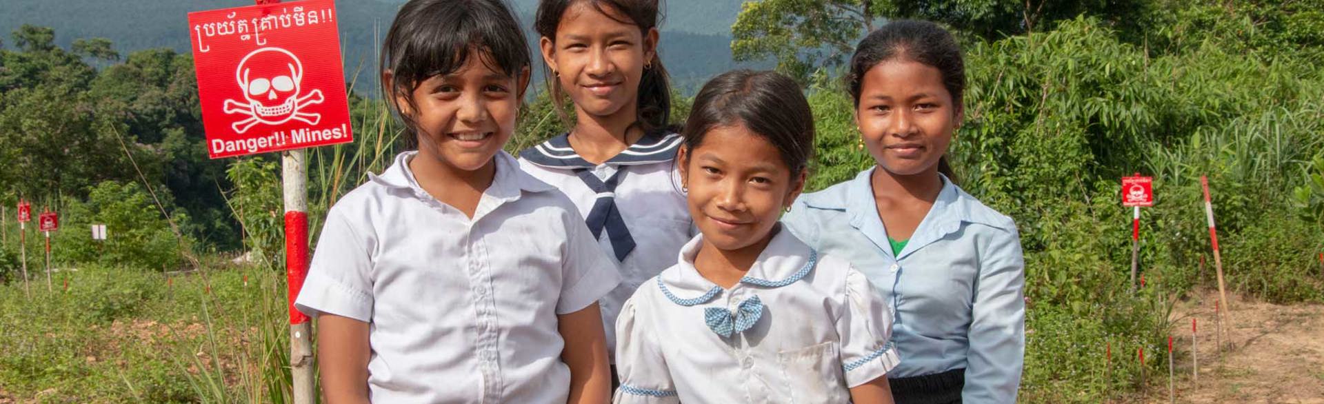 Four school children stand outside in an area marked with mine warning signs