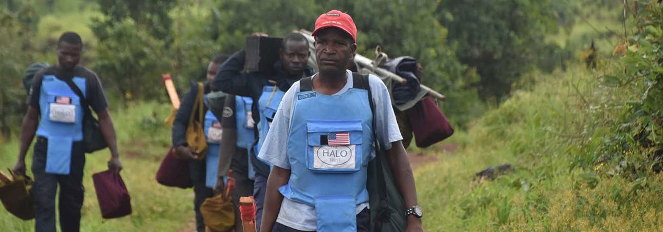 A group of HALO Trust deminers in Angola wearing PPE displaying the US flag carry equipment towards the minefield