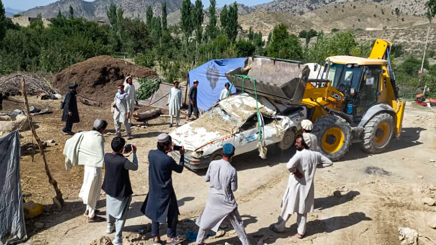 A group of people watch a destroyed car being hauled by a tractor 