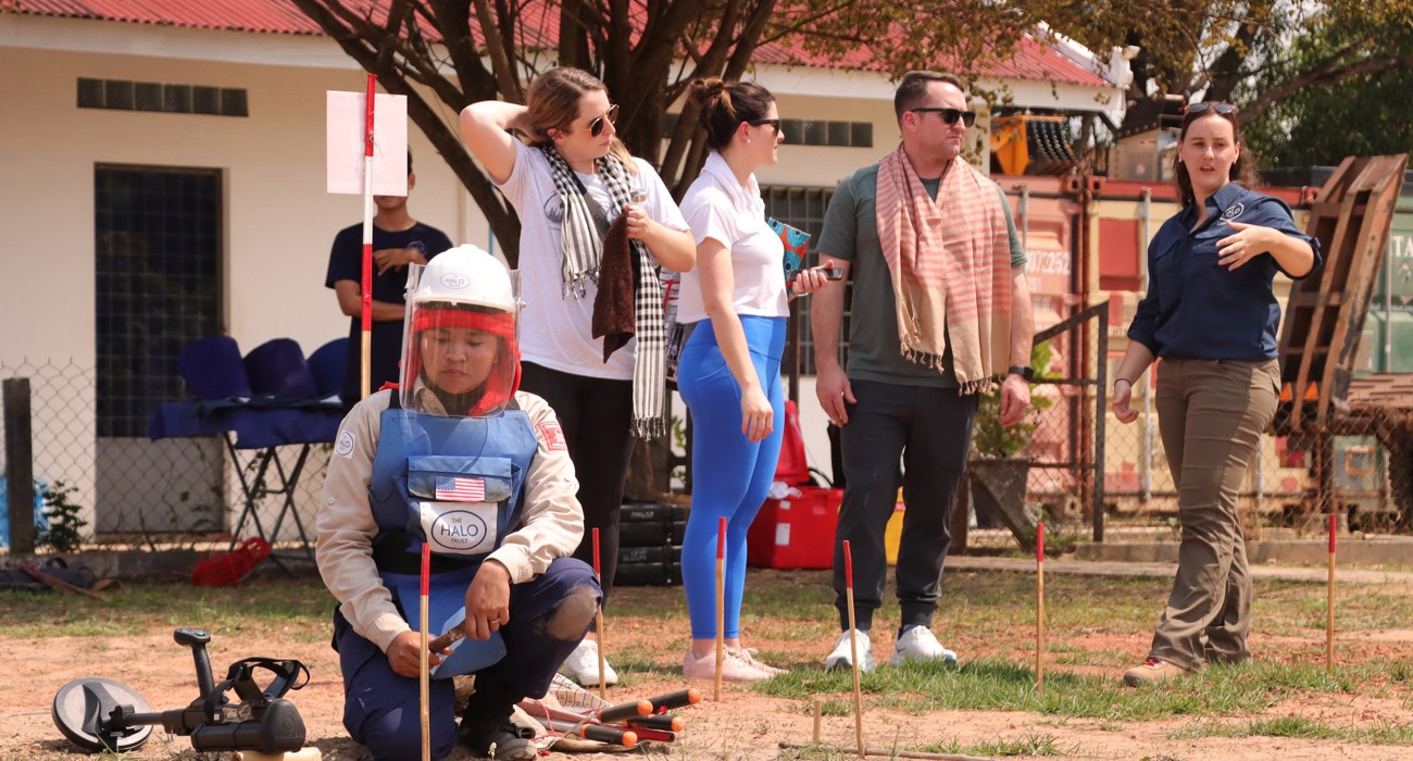 A deminer works on the ground as US Congressional staffers listen to a HALO staff member talking