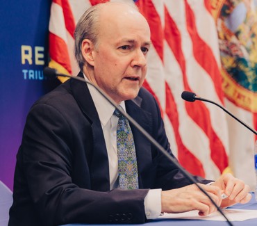 Brian McKeon sits in front of a microphone, an American flag in the background
