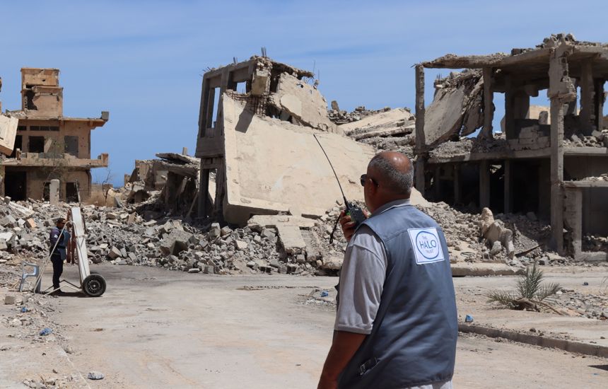 A HALO deminer talks into a radio in front of a collapsed building in Libya