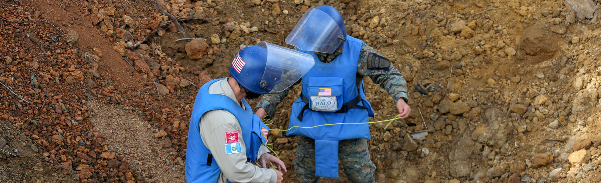 Deminers wearing PPE hold wires as they carry out a bulk demolition on an unexploded ordnance