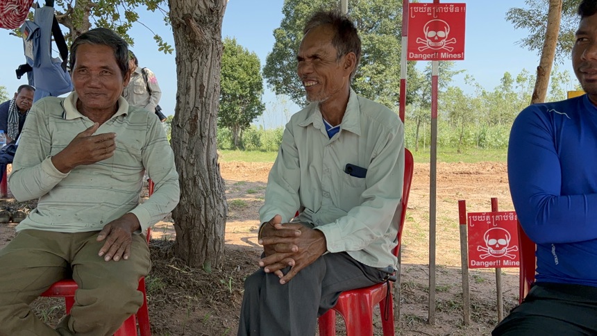 Yul Hai and his neighbour sit on plastic chairs outside with mine warning signs behind them