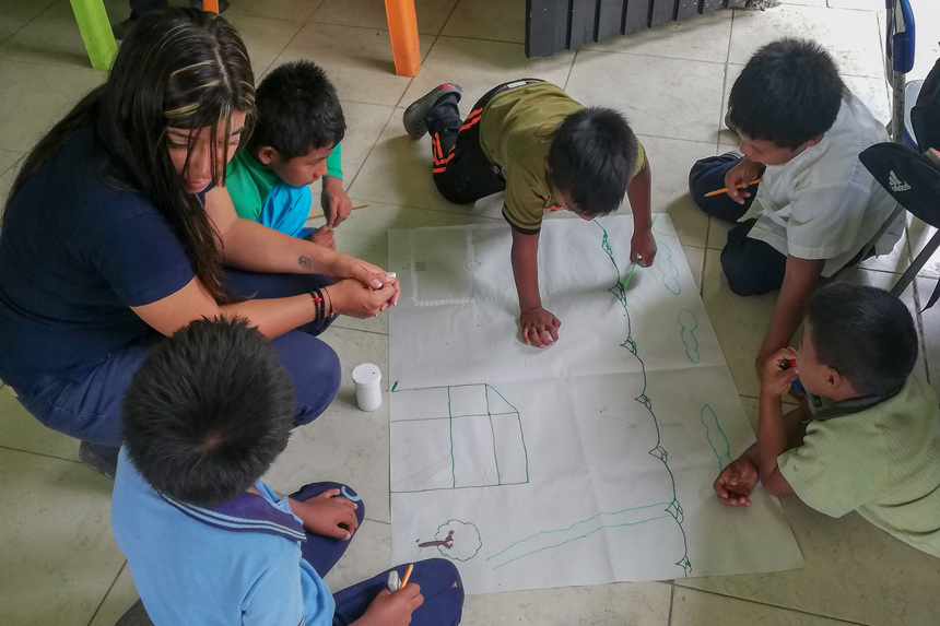 An adult and five children sit on the floor to draw on a large piece of paper