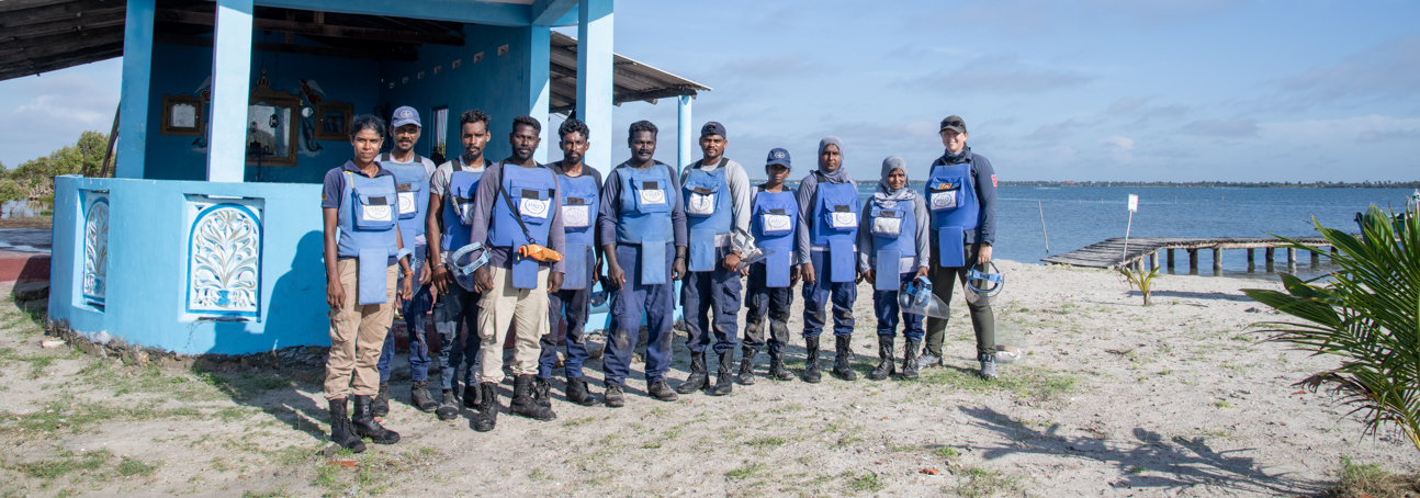 A group of men and women in HALO demining PPE stand in front of a coastal church.