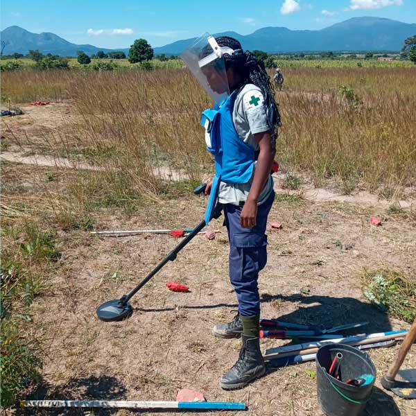 A woman dressed in PPE uses a metal detector to scan the ground for mines
