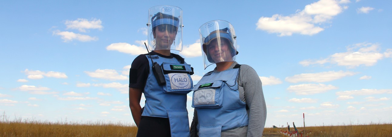 Two deminers in vests and masks stand in a field looking at the camera.