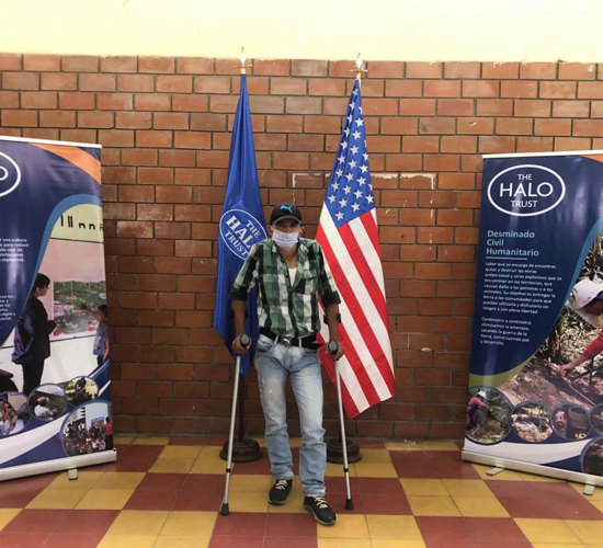A landmine victim stands on crutches with HALO banners and a US flag behind him