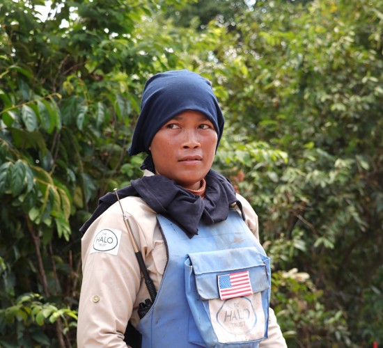 A female deminer stands in a blue vest with an American flag against a green background.