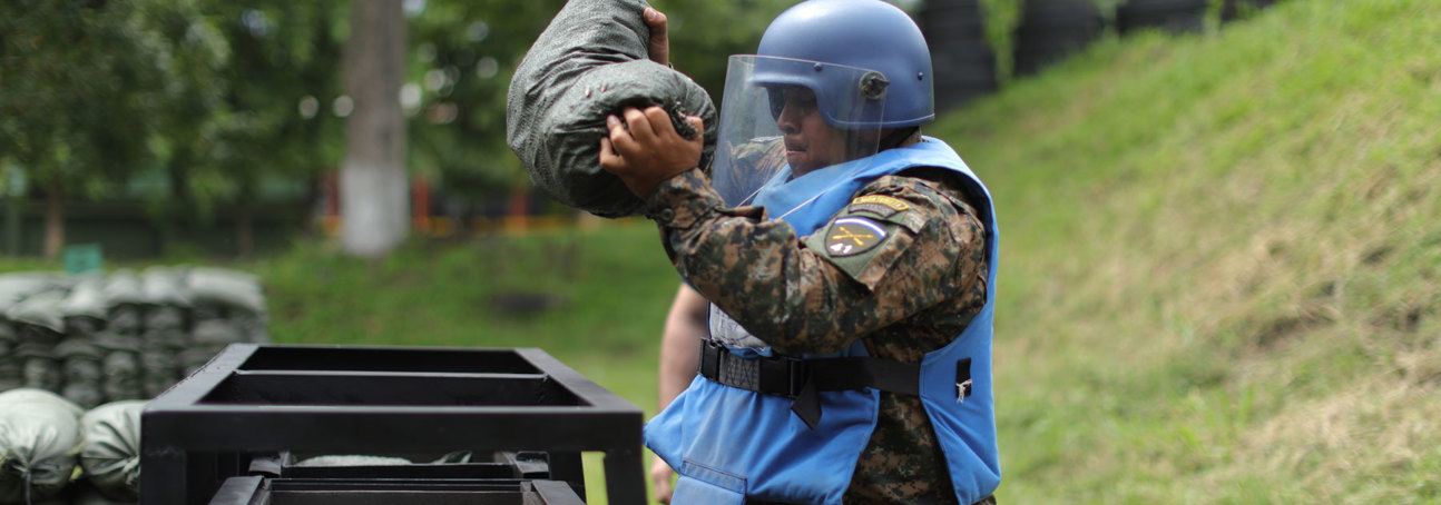 A HALO Trust Weapons and Ammunition technician holds a large sandbag whilst wearing a protective helmet and visor
