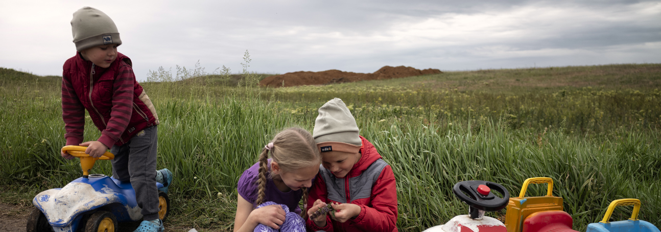 Two young children play together in a field. One boy rides a toy car to the left side.