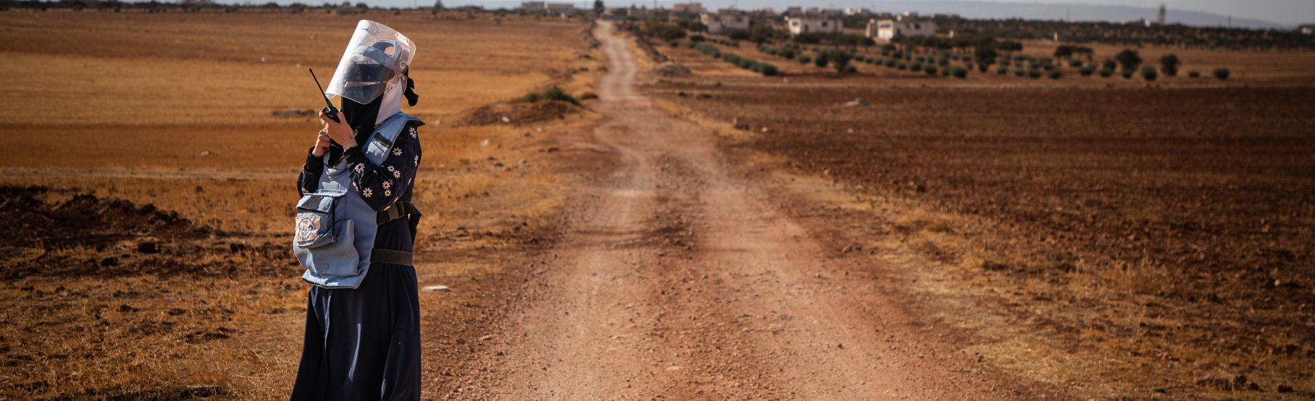 Deminer Hiba talks on a portable radio in front of a long stretch of dirt road.