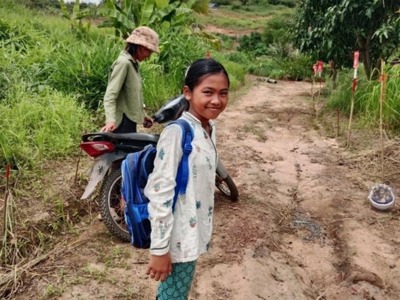 A young girl wearing a backpack stands in front of a path through high grasses marked by minefield signs on one side