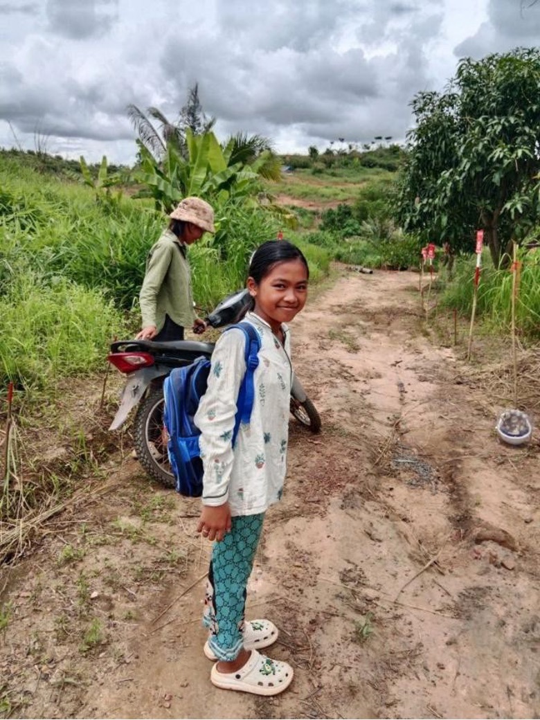 A young girl wearing a backpack stands in front of a path through high grasses marked by minefield signs on one side