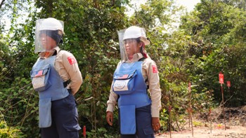 Two Cambodian deminers, wearing US Flags on their PPE vests, stand in a minefield