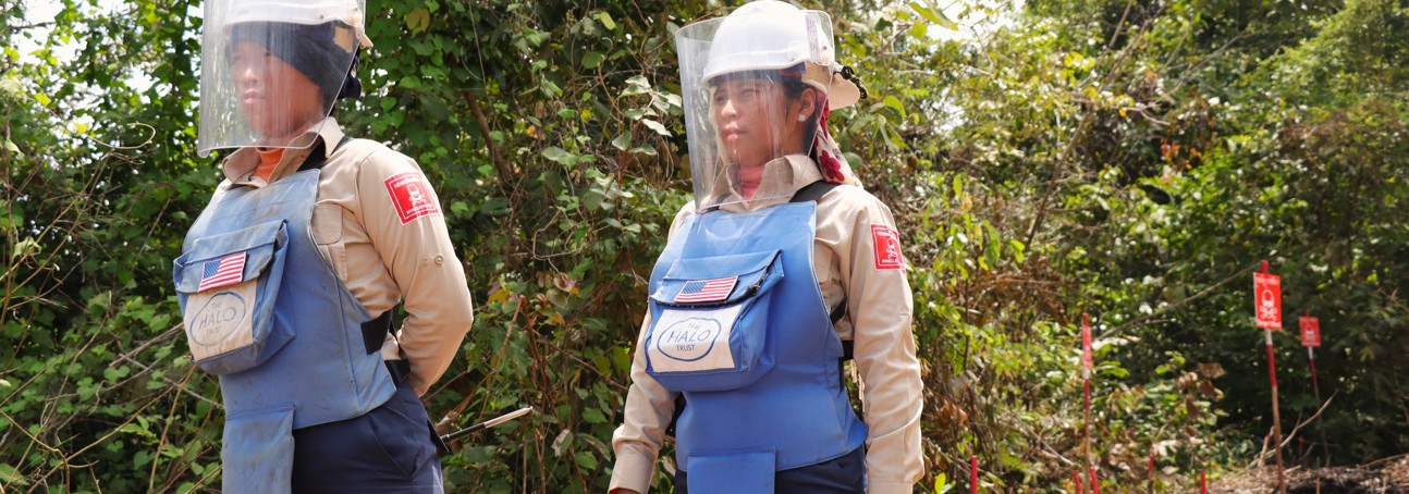 Two Cambodian deminers, wearing US Flags on their PPE vests, stand in a minefield