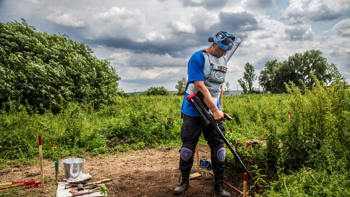 A deminer wearing PPE uses a metal detector to scan the shrubs amongst mine markers