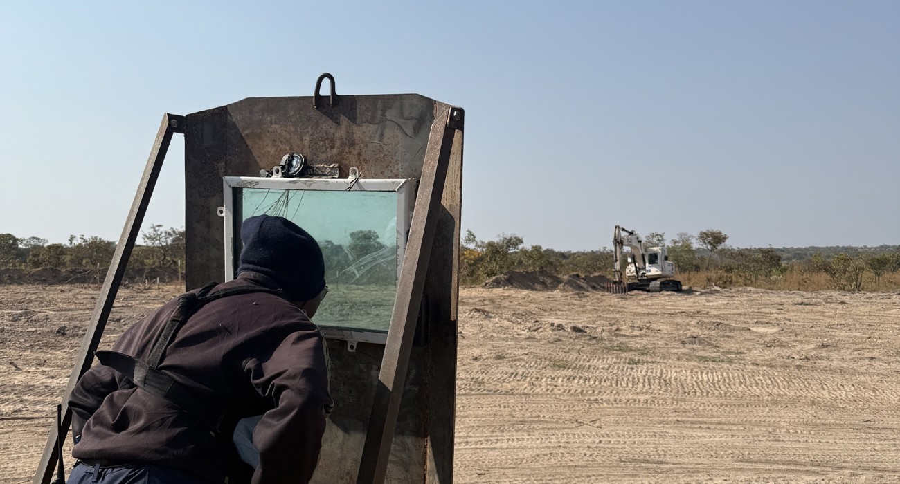 A deminer stands behind a screen observing a mechanical excavation.