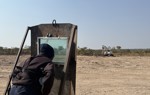 A deminer stands behind a screen observing a mechanical excavation.