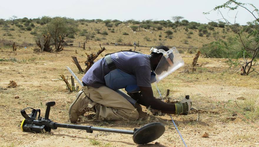 A deminer wearing PPE kneels on the soil to carry out demining activities next to his detector