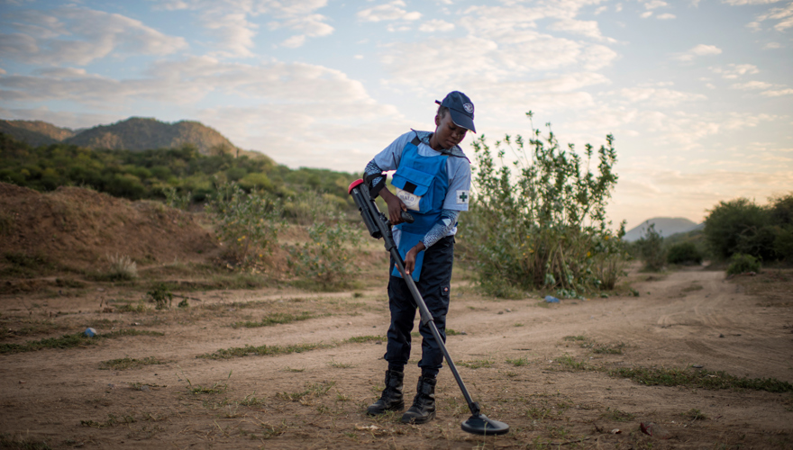 A female deminer holds a metal detector above the soil in order to locate mines