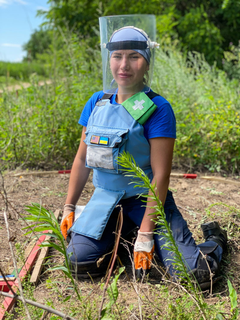A deminer in Ukraine kneeling while wearing a visor and blue PPE