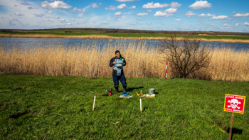 A HALO deminer stands in PPE with his equipment in a grassy minefield in front of a lake