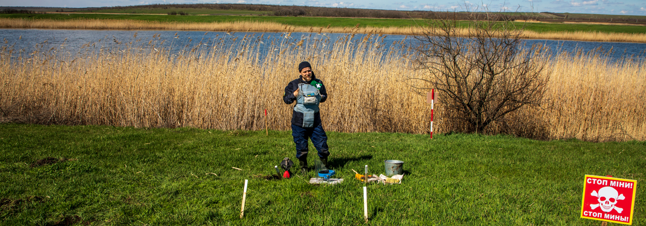 A HALO deminer stands in PPE with his equipment in a grassy minefield in front of a lake