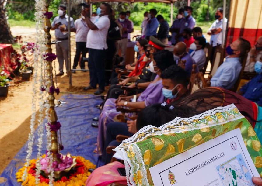 locals sit outside at Muhamalai handover ceremony, a land release certificate can be seen in the corner