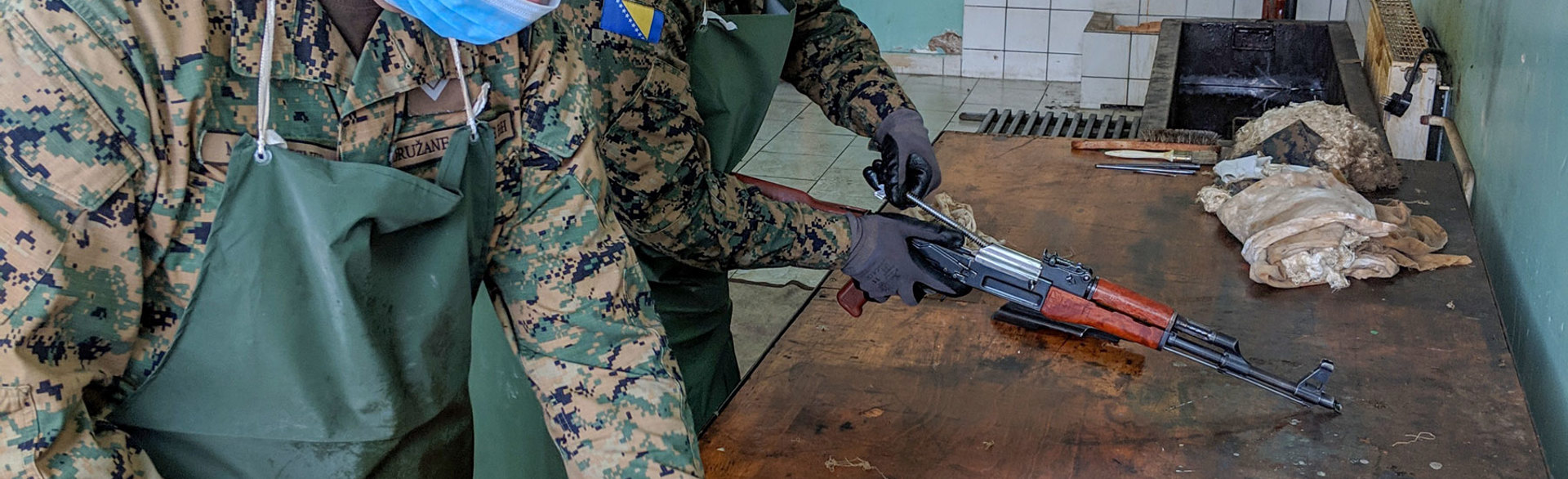 Two men in wearing face masks and gloves clean guns on a work table