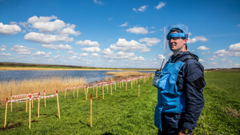 A HALO Trust deminer stands in a rural minefield next to a lake, with contamination clearly marked in front of him