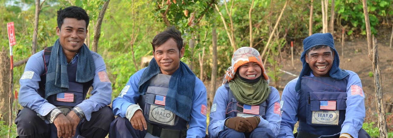 The demining team in Cambodia pose kneeling down and smile whilst wearing their PPE 