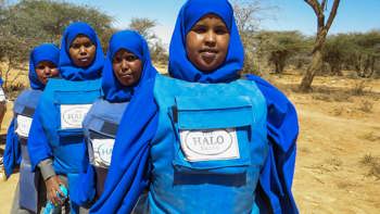 Four female deminers stand lined up in a rural area wearing PPE