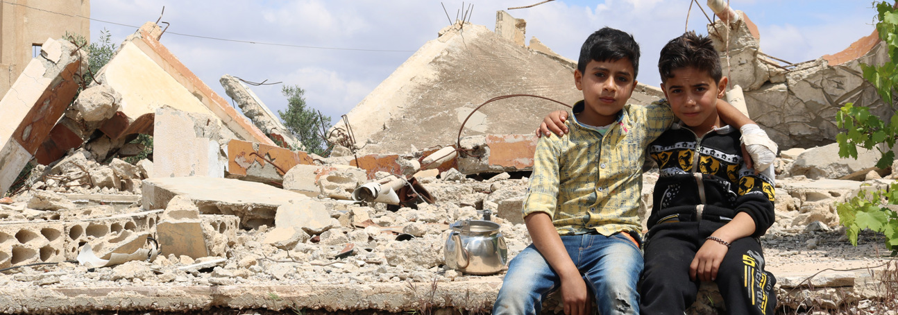 Two young boys sit in front of a destroyed house